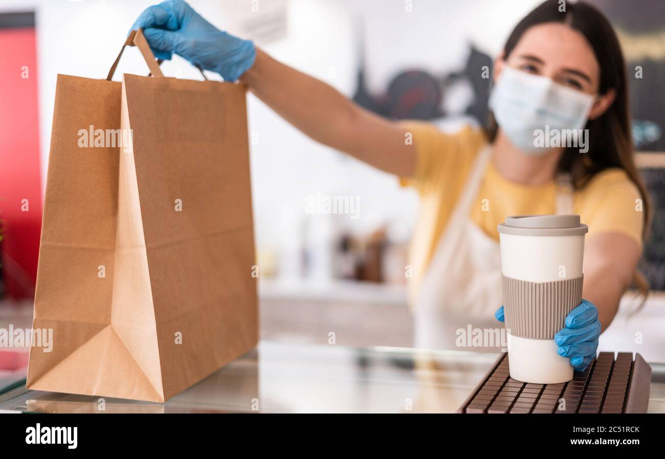 Young woman wearing face mask while serving takeaway breakfast and coffee inside cafeteria restaurant - Worker preparing delivery food inside bakery b Stock Photo