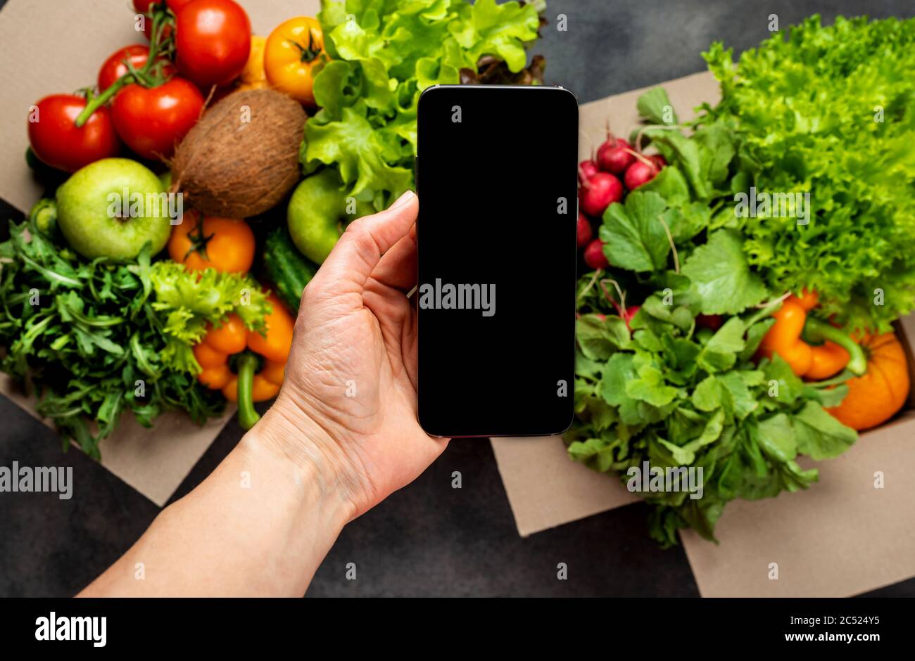 Female hand with smartphone and full boxes of different fresh organic vegetables. Delivery or Order food online concept. Stock Photo