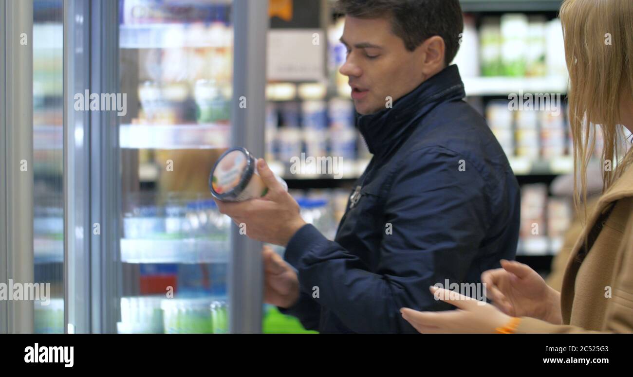 Young couple shopping in the supermarket. Man taking ice-cream from the fridge, but woman is angry with him. He putting it back, but returning alone a Stock Photo