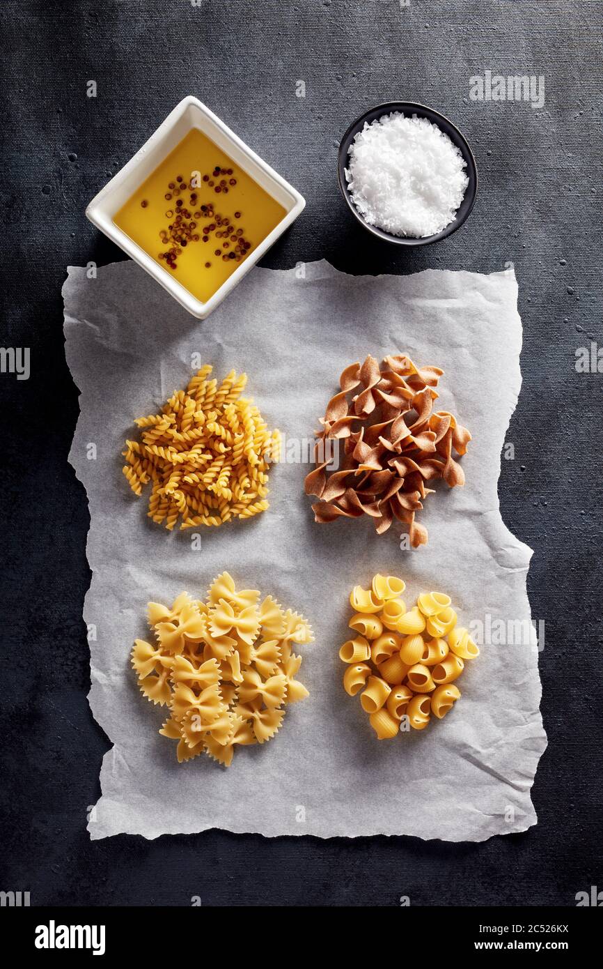 Variety of raw pasta with olive oil and salt on baking paper on black background. Flat lay view. Stock Photo