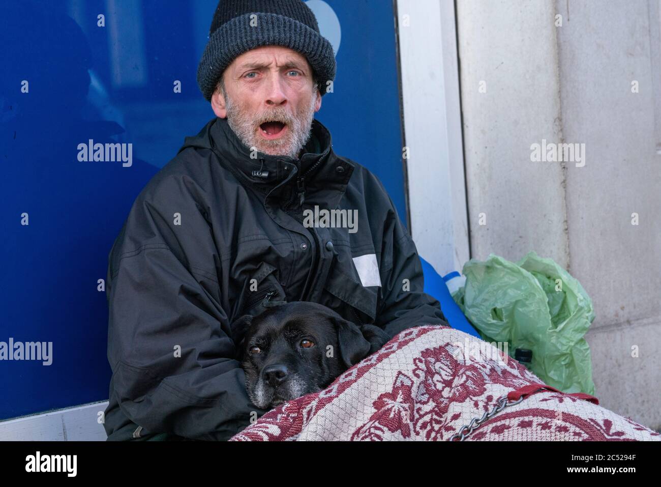 An elderly homeless man, sits with his back against a shop window. Cuddling a dog, while shouting at people who pass by. Stock Photo