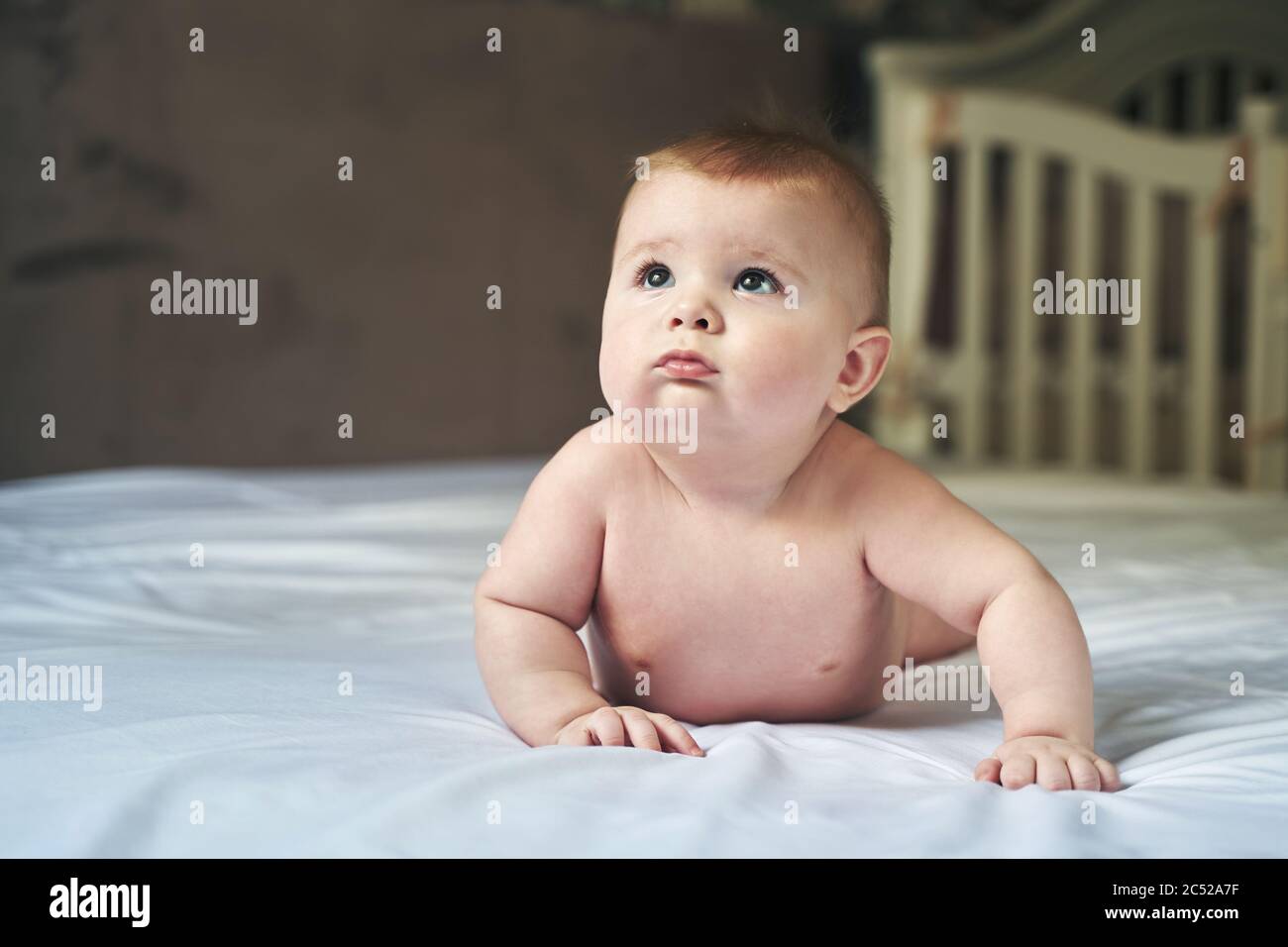 a wonderful six month old baby lies on a large bed on a white sheet and looks up Stock Photo
