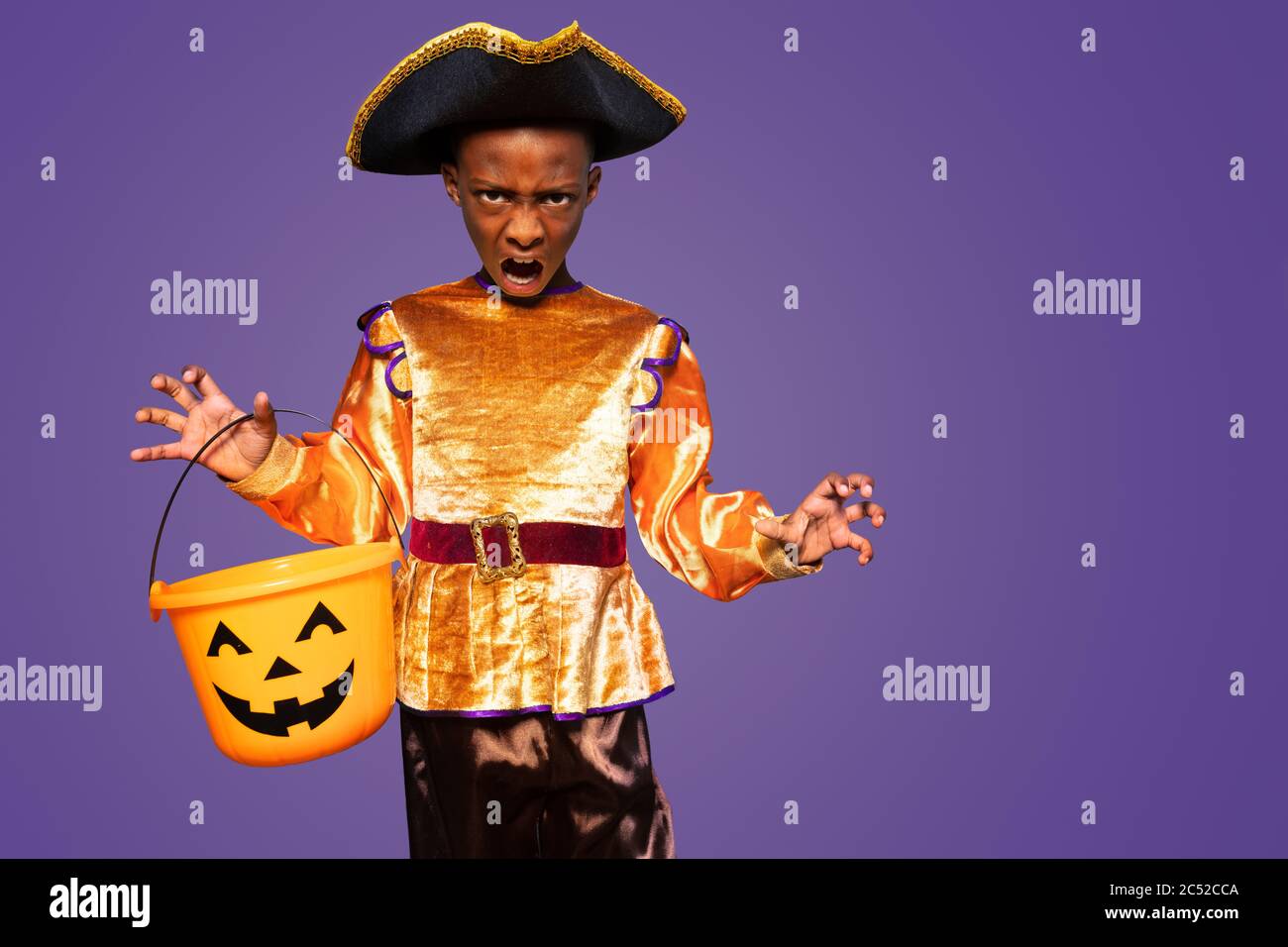 Little boy in Halloween costume show spooky gestures standing over purple background holding scary candy bucket Stock Photo