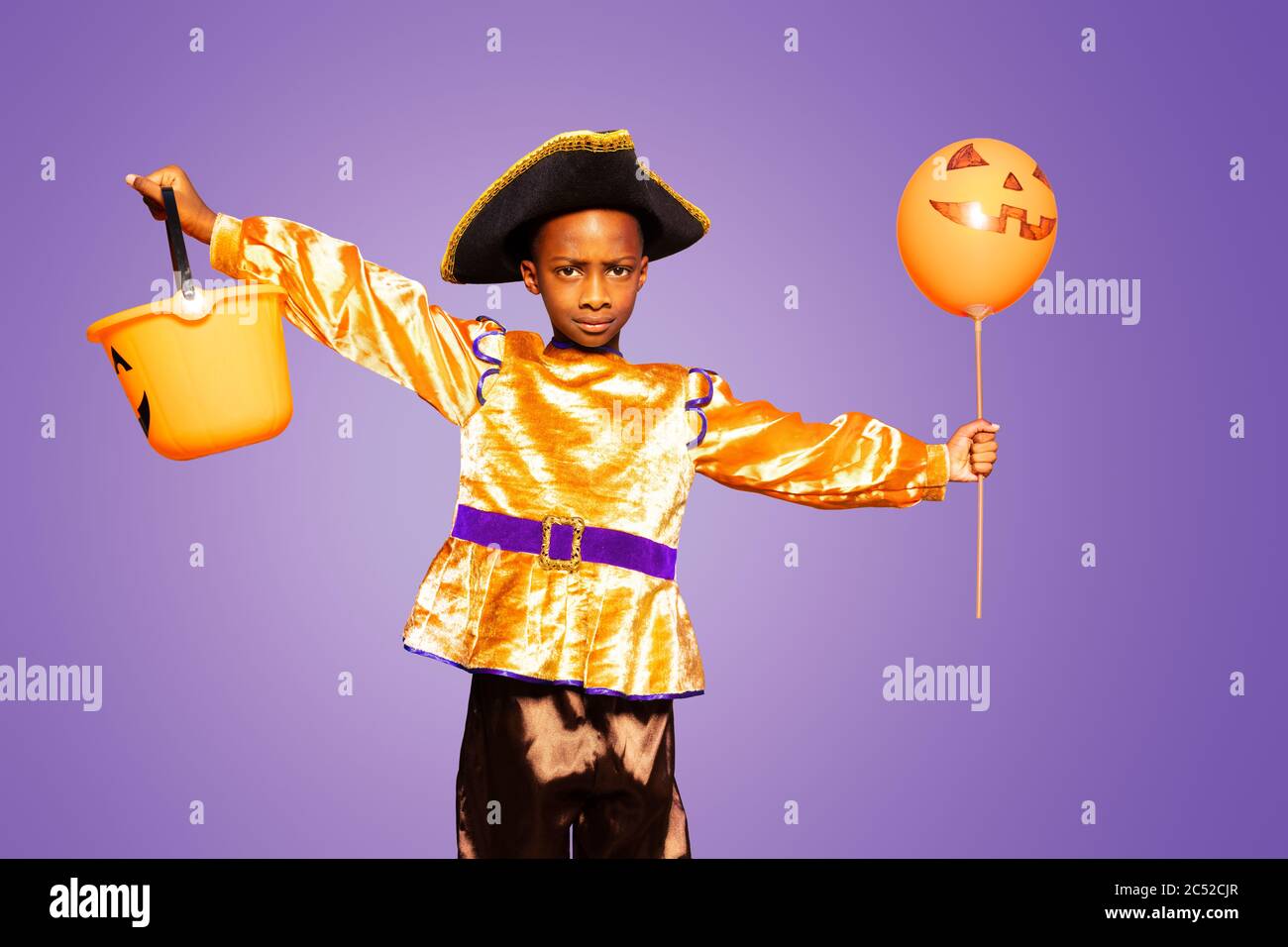 Little cute black boy in Halloween costume spread hands with serious face standing over purple background holding scary candy bucket Stock Photo