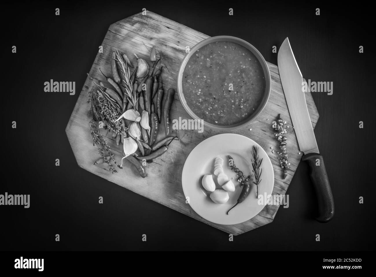 Hot Red Chilli sauce served in a blue bowl, with chili’s, garlic, and herbs being prepared, on a black textured background Stock Photo