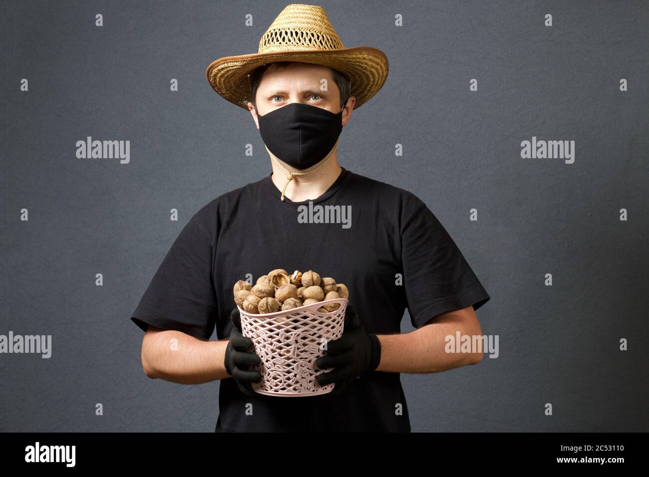 A farmer in a straw hat and a protective mask holds in his hands a wicker basket with walnuts. On a gray background. Harvest during the 2020 pandemic. Stock Photo