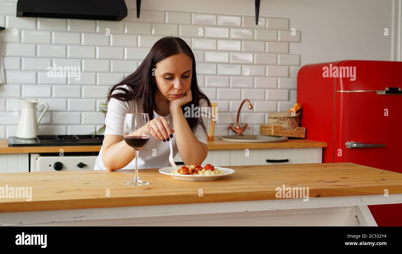 Young woman picks with fork in dish. Adult female sits thoughtfully in kitchen over plate of food. Stock Photo