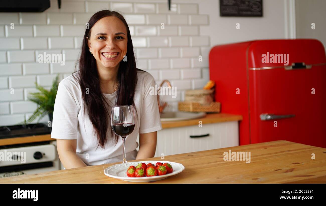 Young woman drinks red wine and eats strawberry, sitting at kitchen table. Adult alone female relaxes at home. Stock Photo