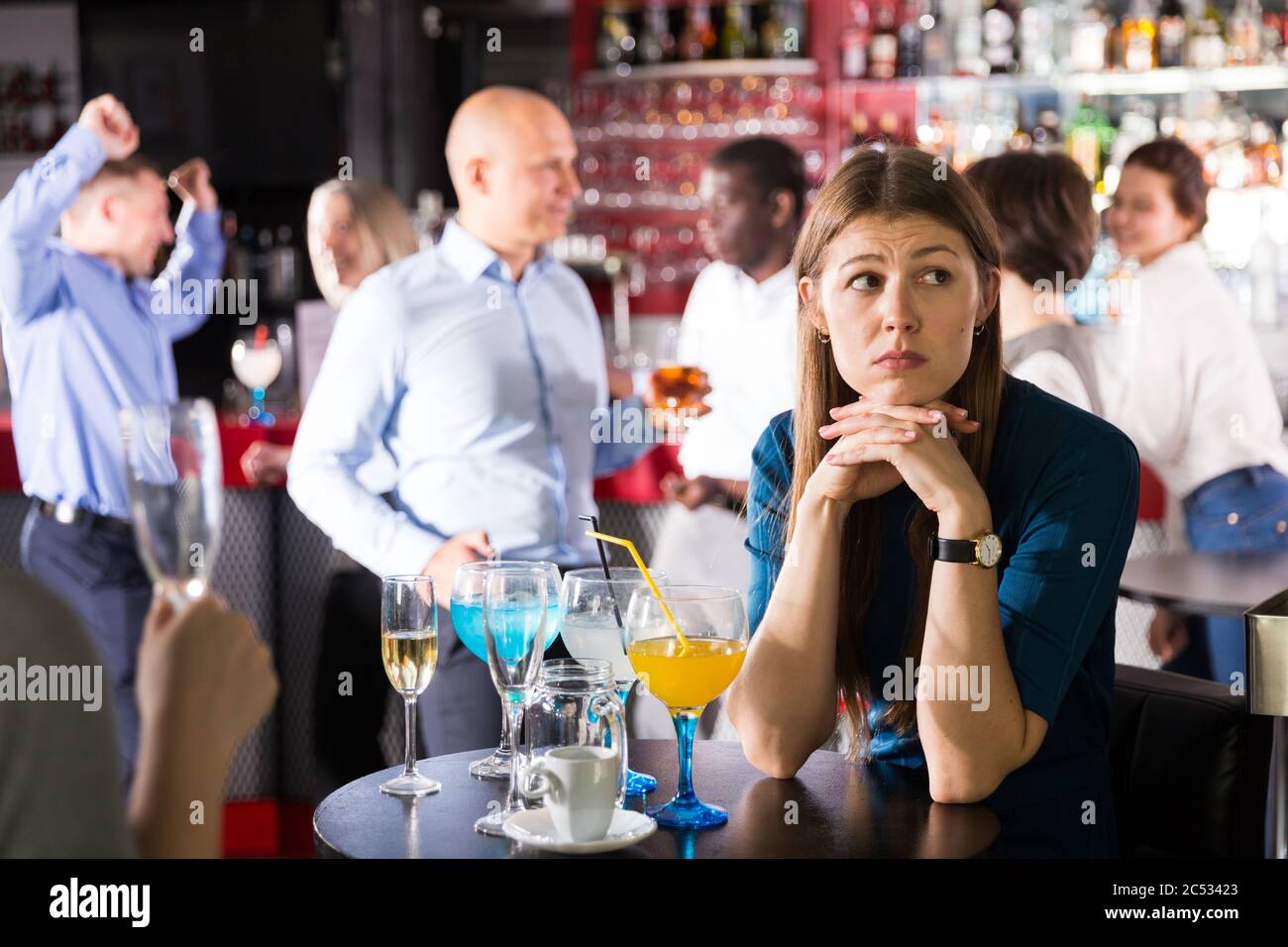 Upset young woman sitting alone at corporate bar party on background with  cheerful colleagues Stock Photo - Alamy