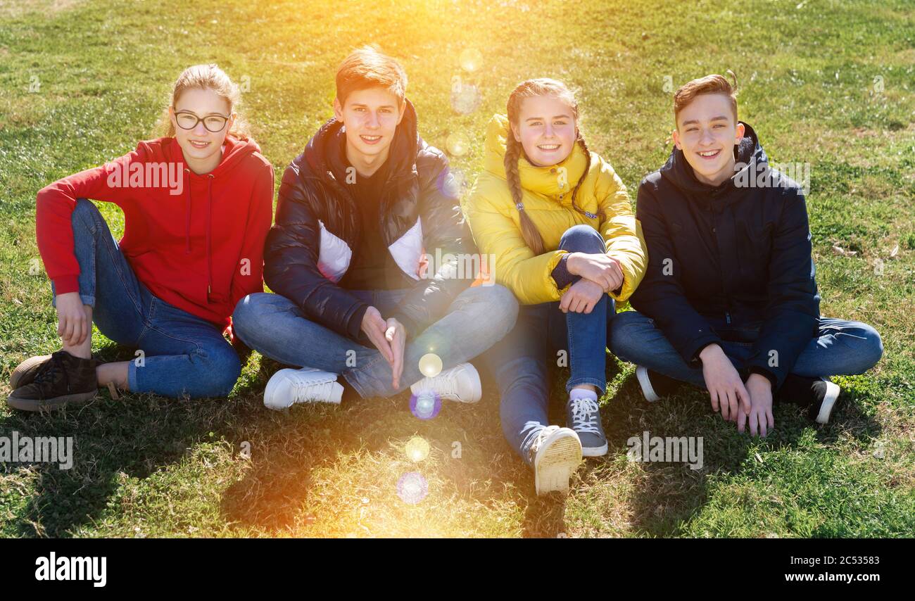 Portrait of four happy teenagers sitting on green lawn of city park on ...