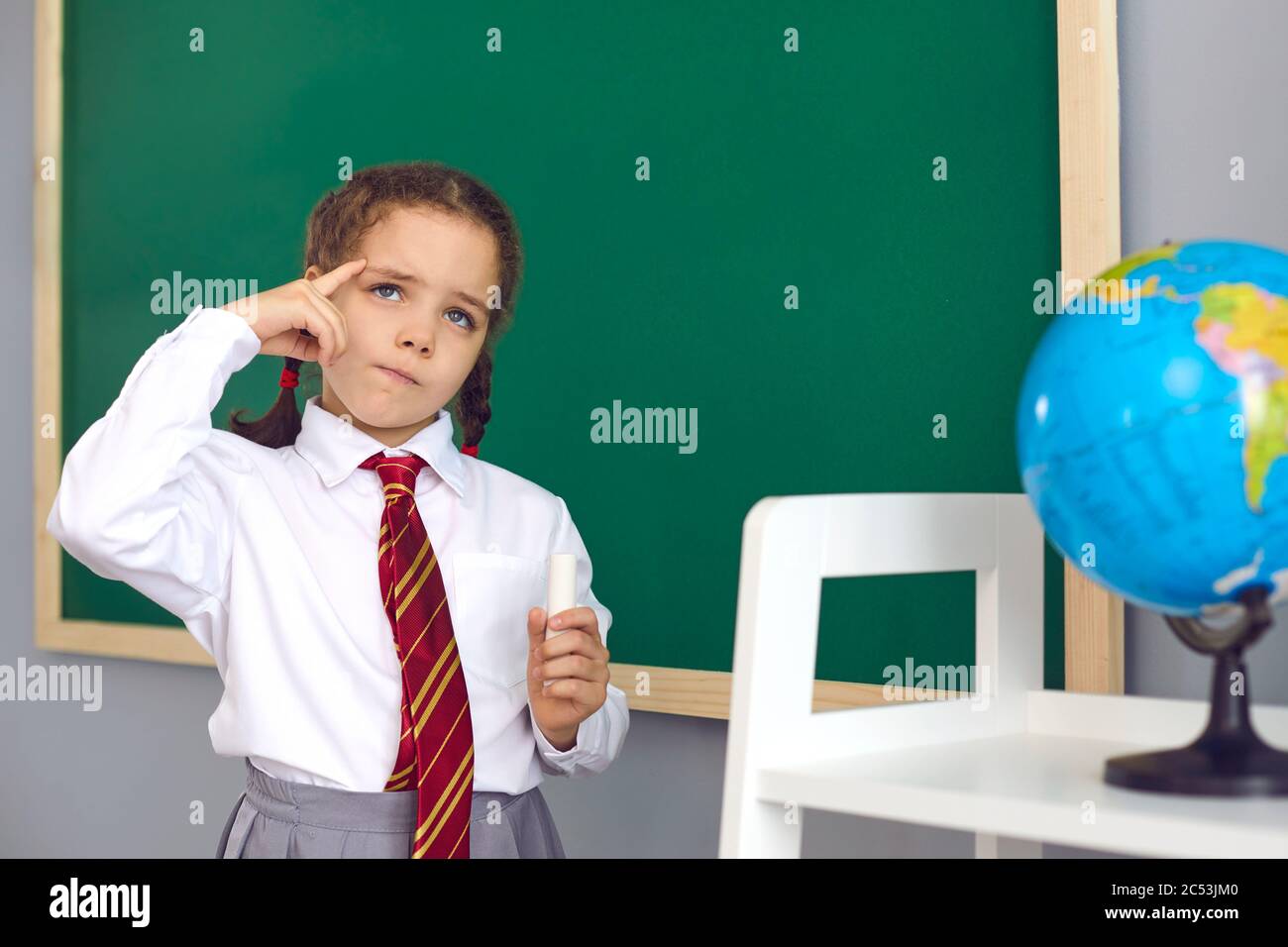 Back to school. A happy schoolgirl thinks about the task while standing at the blackboard in the classroom. Stock Photo
