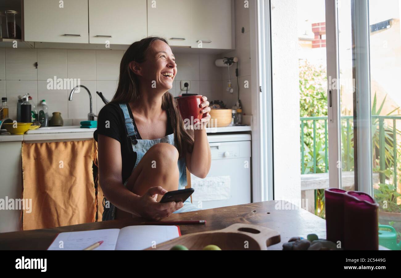 Young woman sitting on a wooden kitchen table with relaxed atmosphere checking her smartphone and writing notes; concept natural lifestyle, simplicity Stock Photo