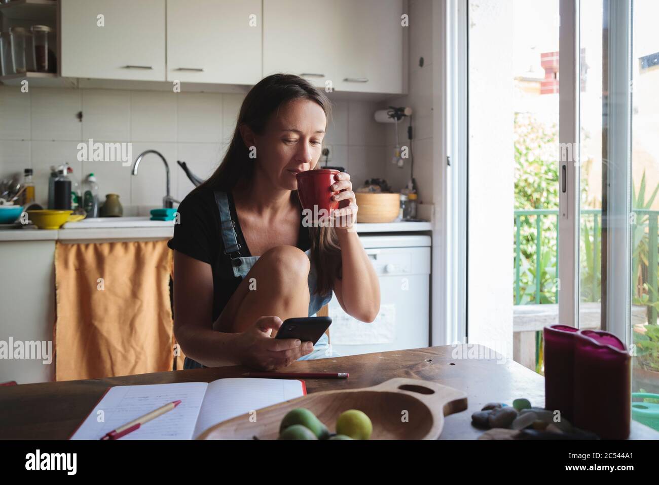Young woman sitting on a wooden kitchen table with relaxed atmosphere checking her smartphone and writing notes; concept natural lifestyle, simplicity Stock Photo