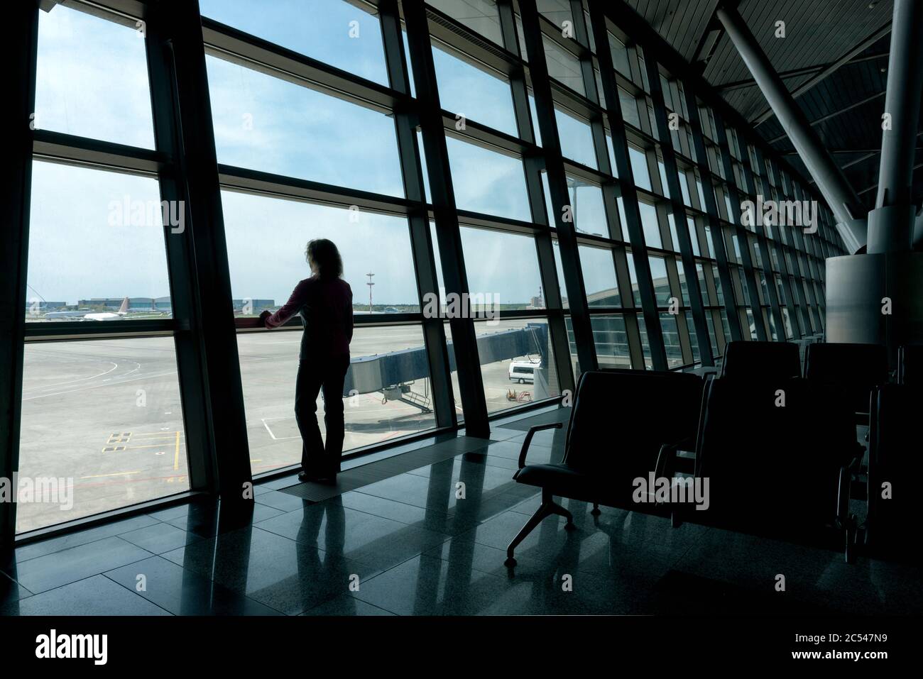 Female traveler looks out the window at the airport terminal. Young woman is waiting for boarding in the hall before departure. Stock Photo