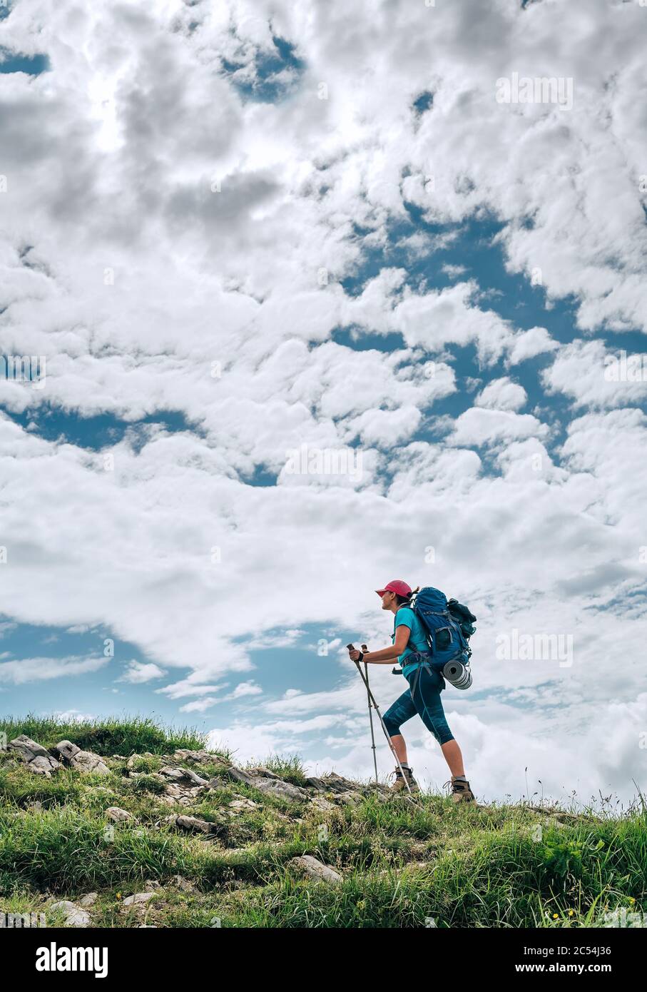 Vertical image of female backpacker climbing to the mountain top using trekking poles with bright cloudscape background. Active vacation spending by s Stock Photo