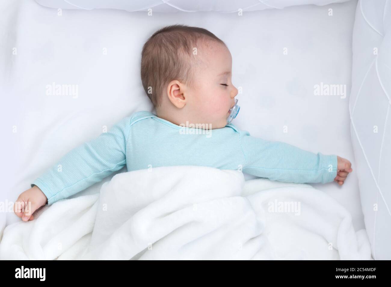 Baby boy sleeping with open arms in a cradle . Light blue pajama and white bed sheets. A pacifier in his mouth. Stock Photo