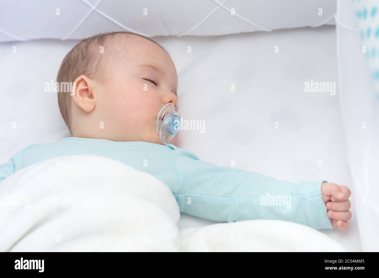 Baby boy sleeping with open arms in a cradle . Light blue pajama and white bed sheets. A pacifier in his mouth. Stock Photo