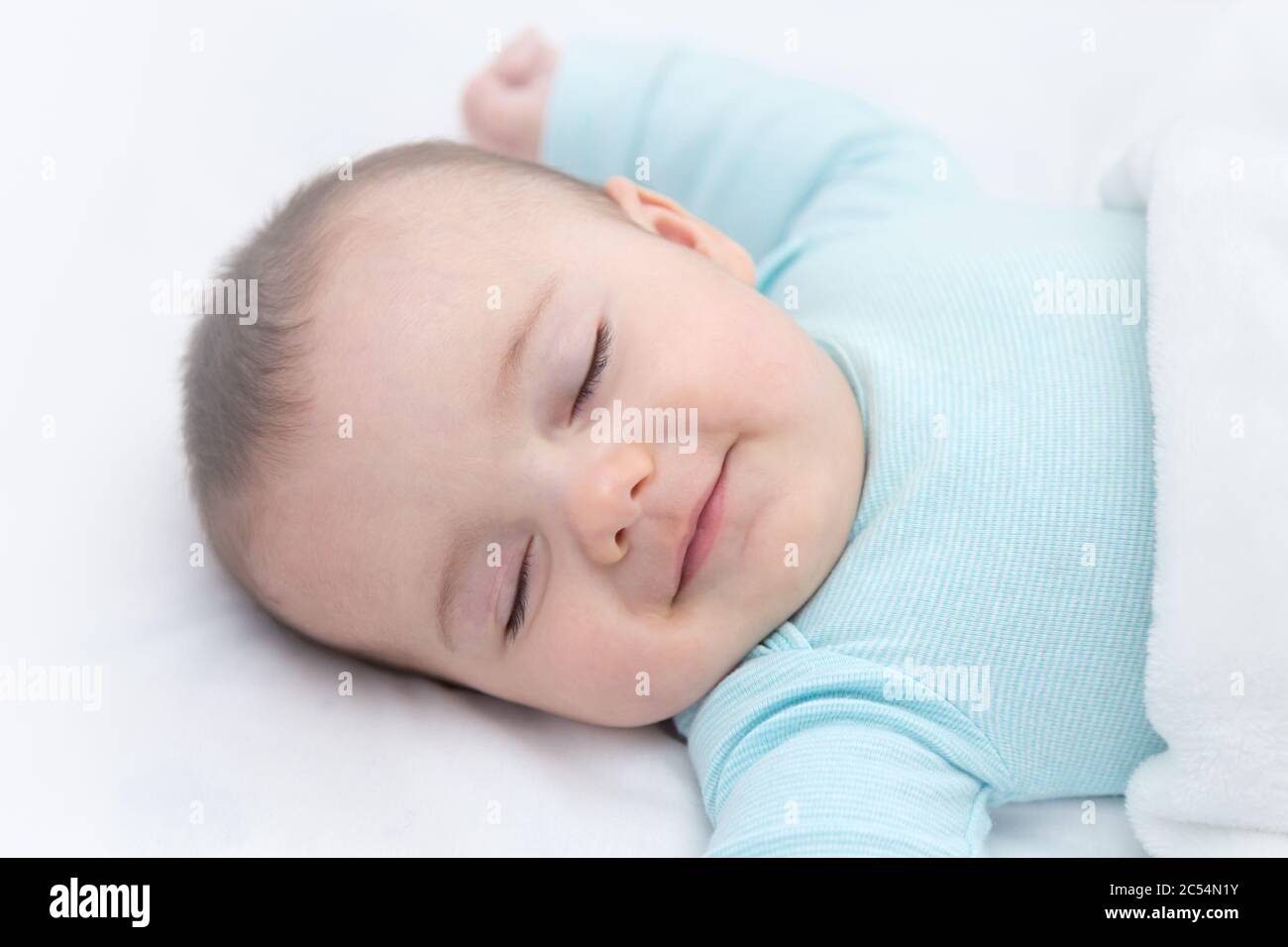 Happy baby sleeping and smiling in a cradle. Light blue pajama and white bed sheets. Stock Photo