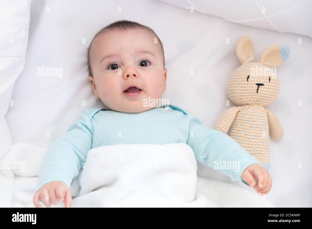 Happy baby newly awake in his crib and with his bunny doll. Light blue pajama and white bed sheets. Stock Photo