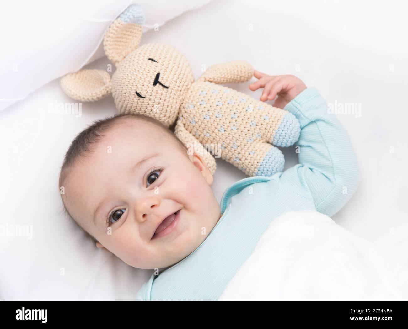 Happy baby newly awake in his crib and with his bunny doll. Light blue pajama and white bed sheets. Stock Photo