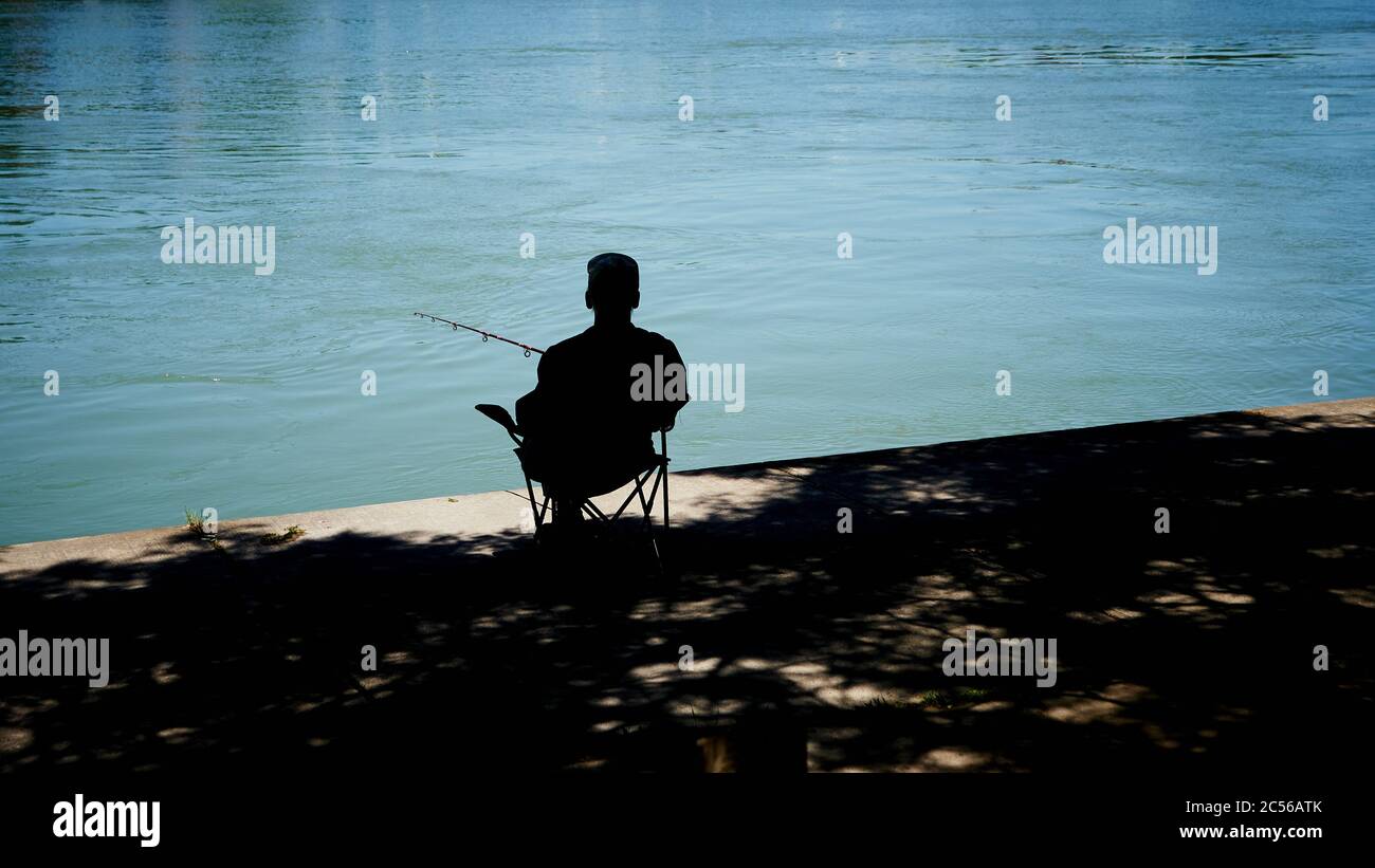 Senior Male in silhouette sits in the shade fishing from a dock at Lake