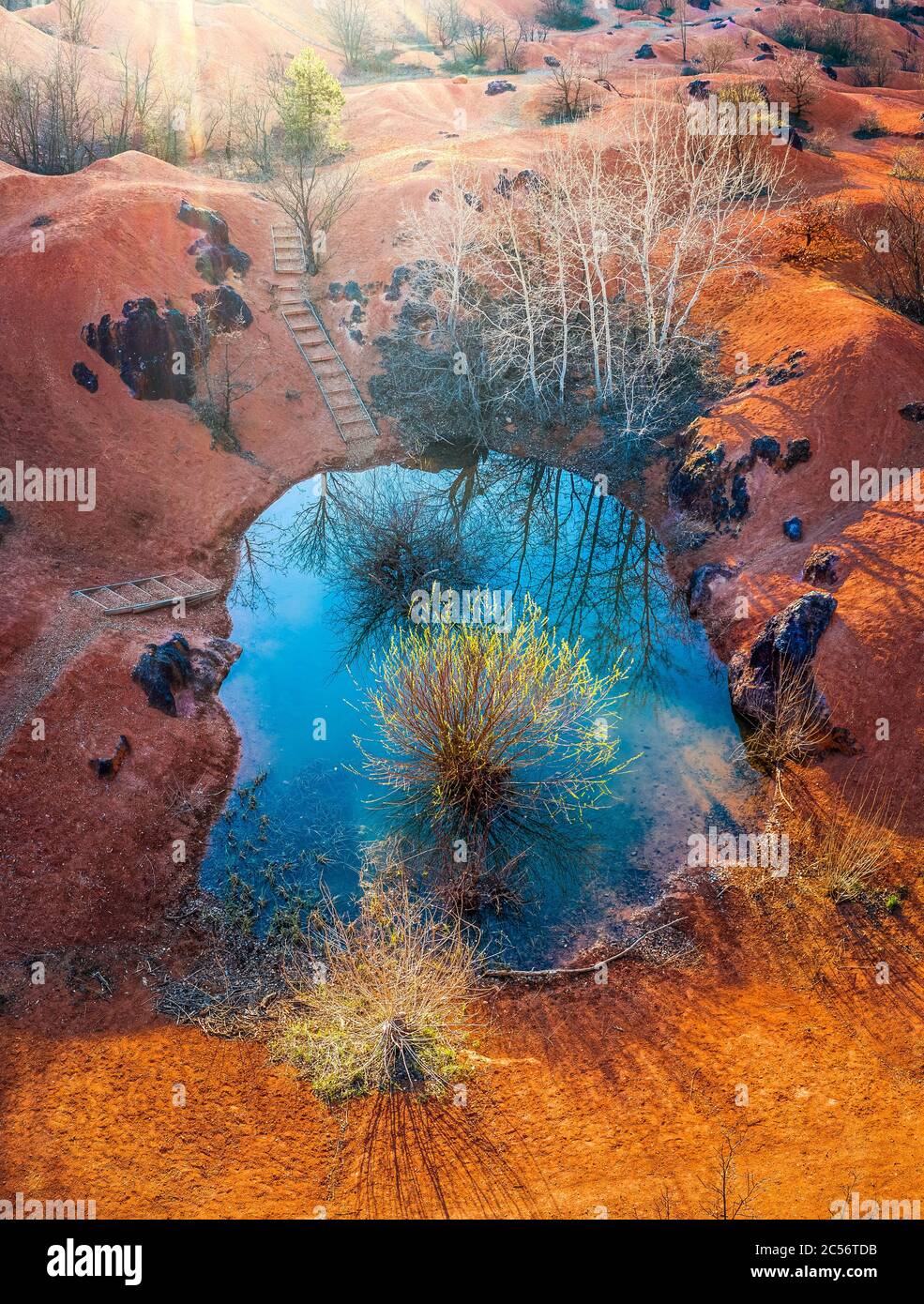 Gant, Hungary - Aerial view of abandoned bauxite mine with small blue lake and warm red and orange colors and tree at sunset Stock Photo