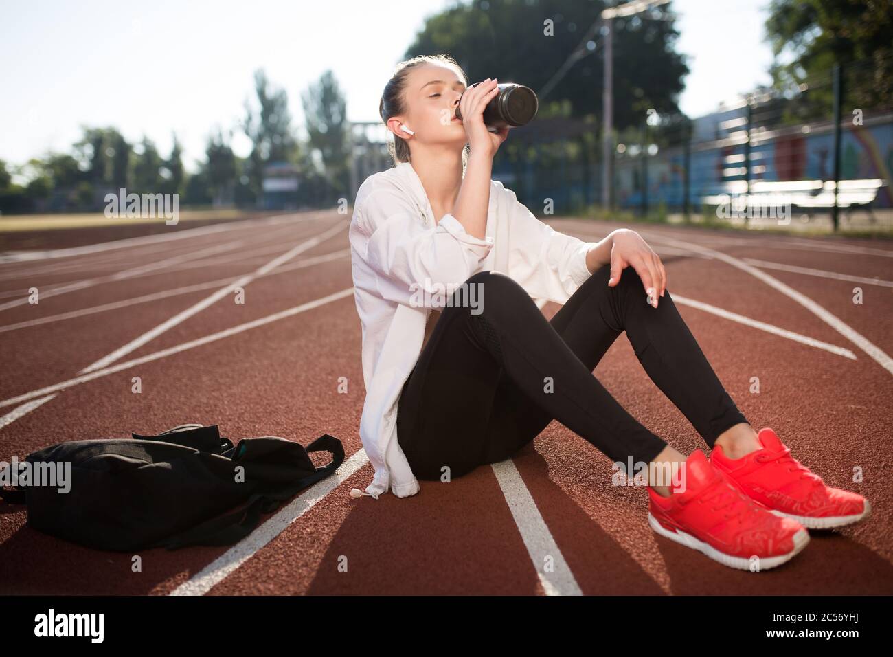 Beautiful girl in wireless earphones drinking pure water while spending time on running track of stadium Stock Photo