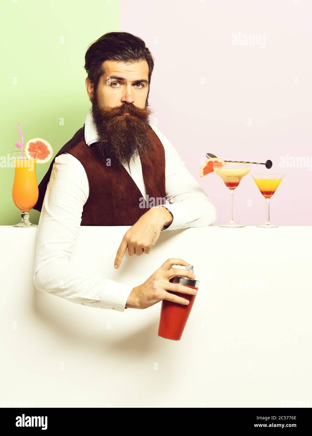 handsome bearded barman with long beard and mustache has stylish hair on serious face holding shaker and made alcoholic cocktail in vintage suede leather waistcoat, on purple green studio background Stock Photo
