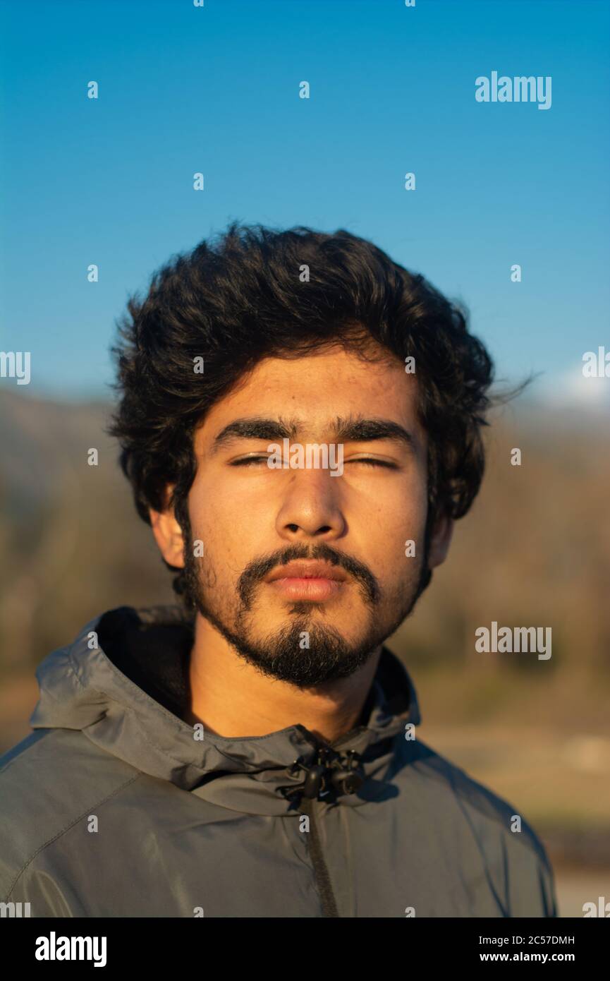 headshot of a handsome 18-20 years old man with his eyes closed in a winter evening/morning. background contains trees, mountains and clear blue sky. Stock Photo