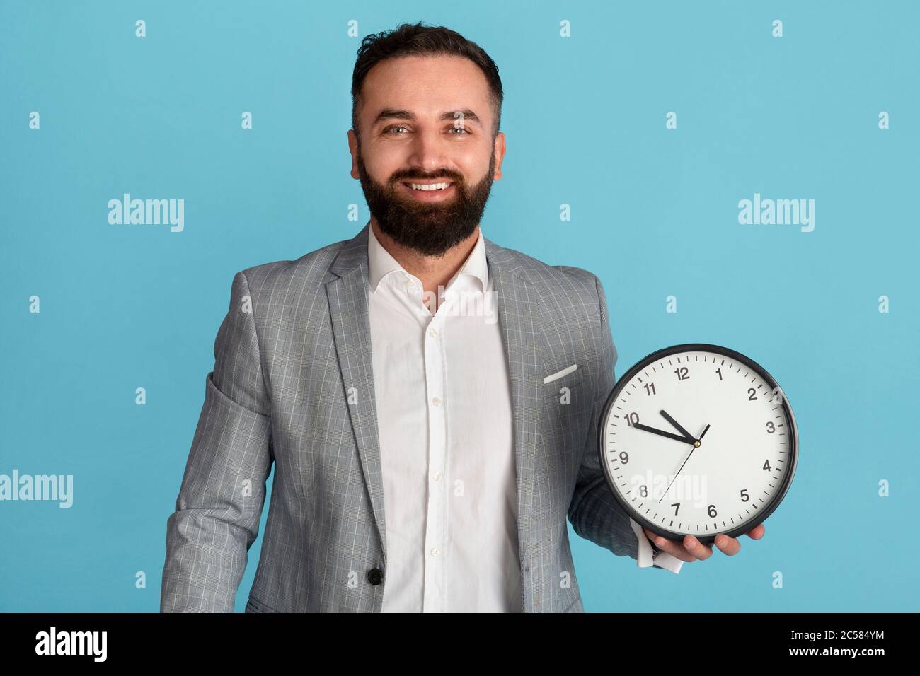 Time management and optimization. Happy businessman holding analog clock on blue background Stock Photo