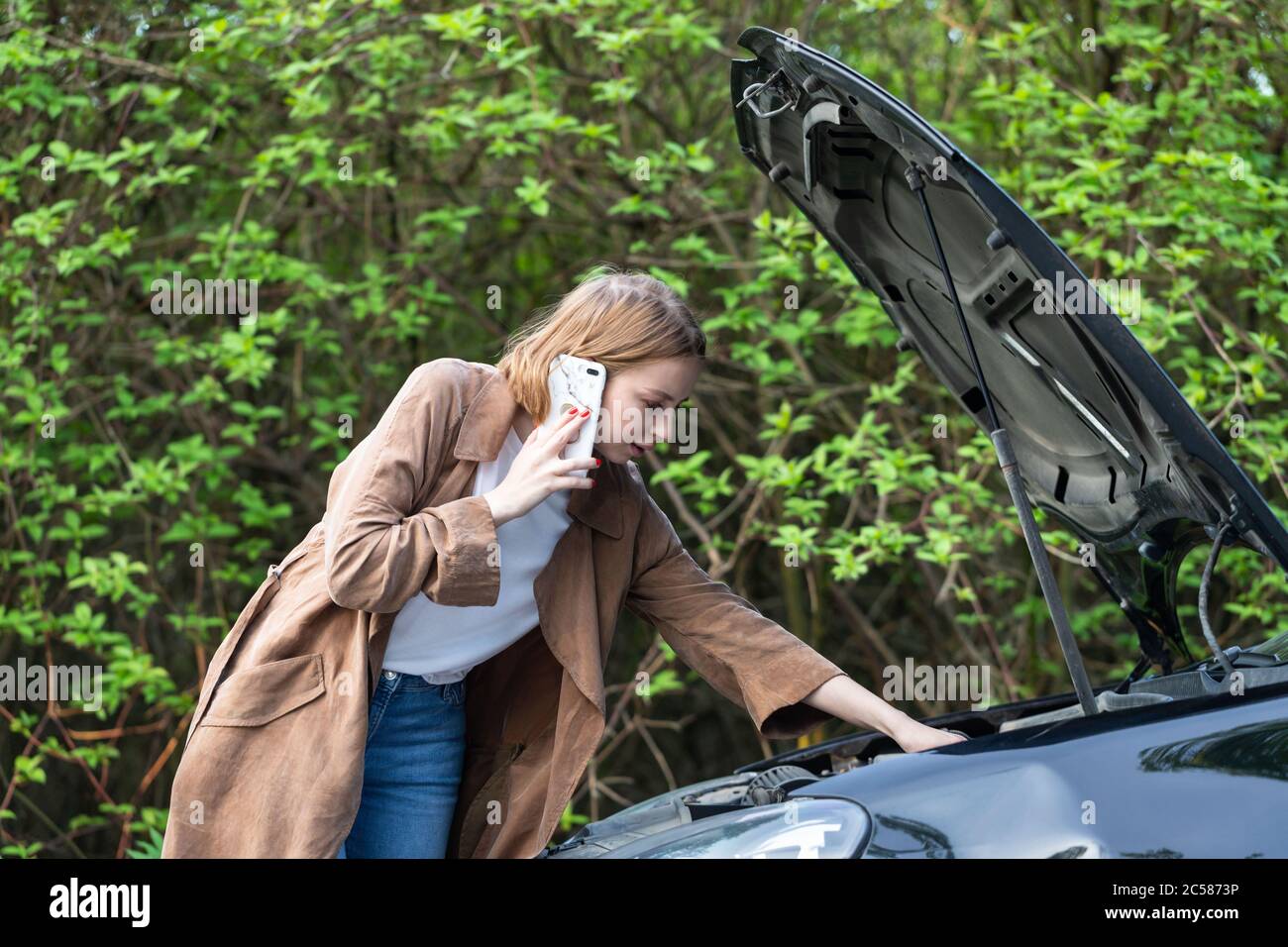 Helpless woman driver calling for help/assistance looking at broken down car, stopped at the roadside. Doesn't understand what happened. Stock Photo