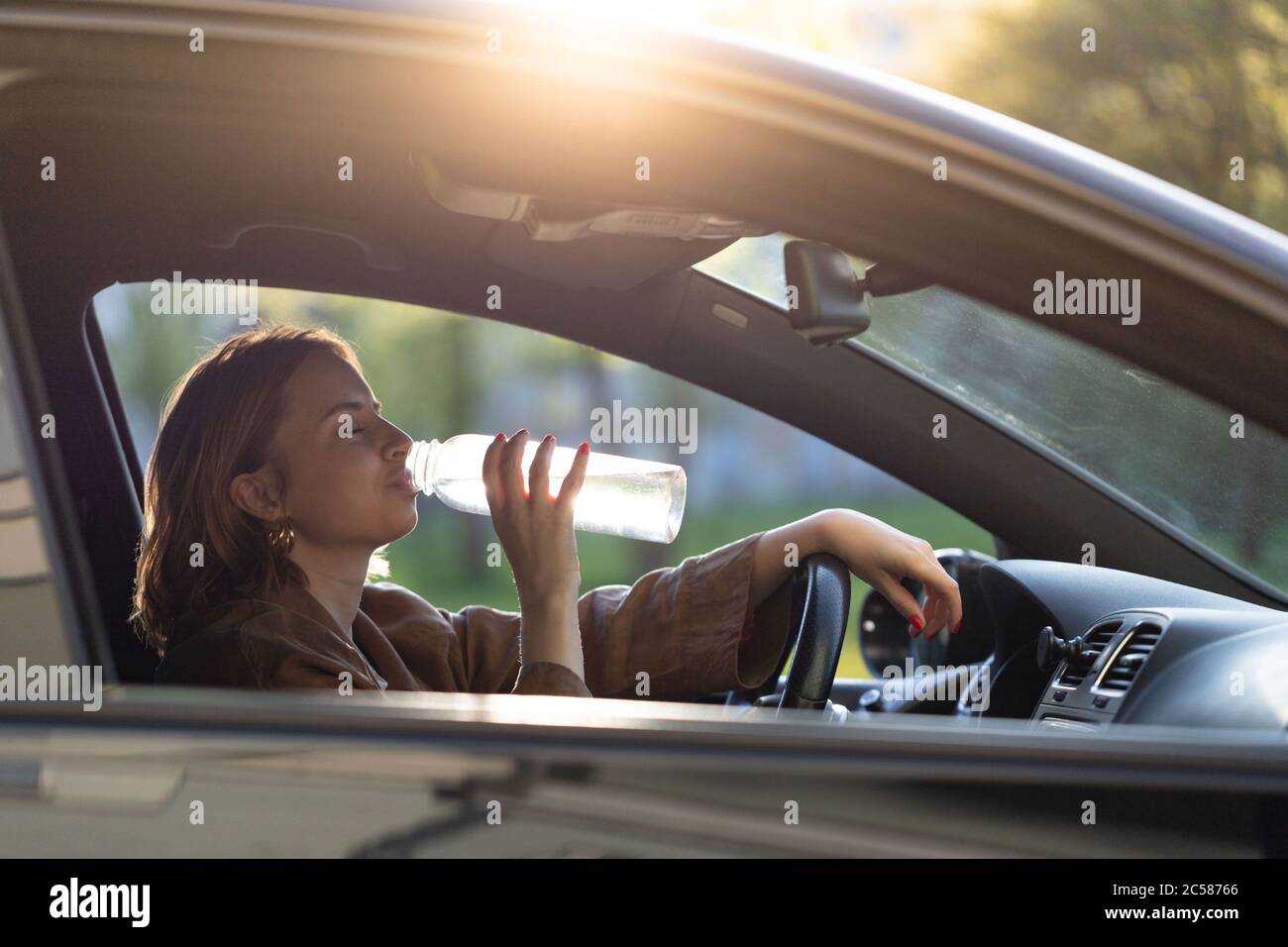 Woman drinks water from a refillable bottle in her car, thirsty behind the wheel, stopped to rest. Stock Photo