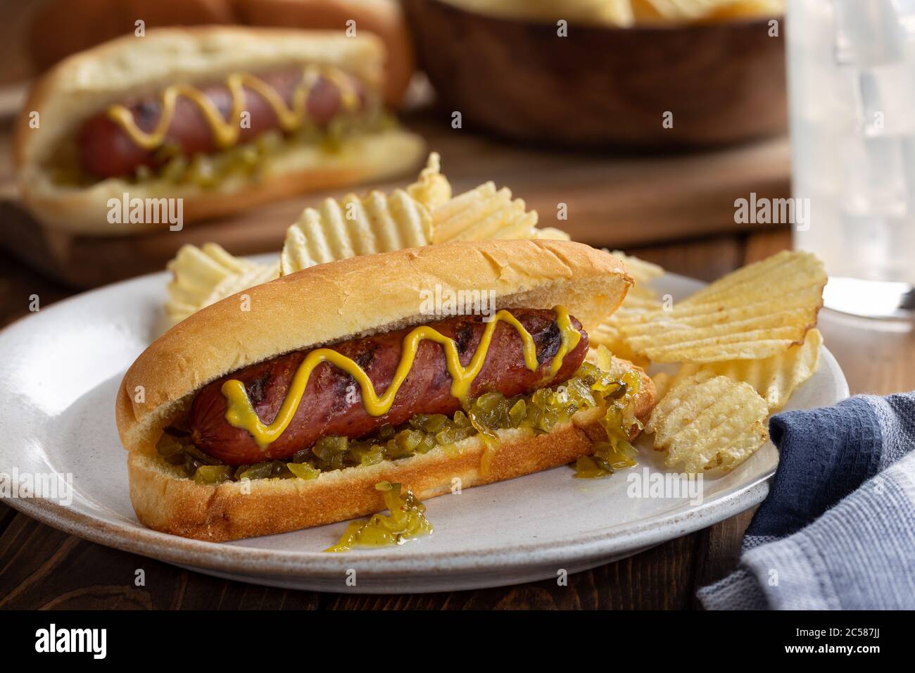 Grilled hot dog with mustard and relish on a bun and potato chips on a white plate Stock Photo