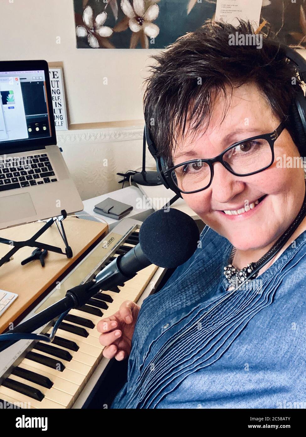 Mature female with black short hair & glasses smiling behind microphone, musical keyboard and computer whilst teaching online through livestreaming Stock Photo