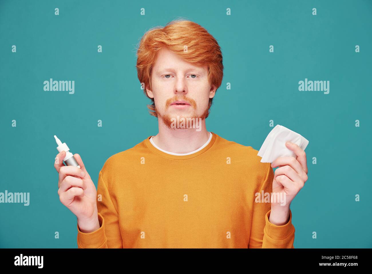 Allergic young man with red beard having stuffy noses and using nasal spray and napkin, blue background Stock Photo