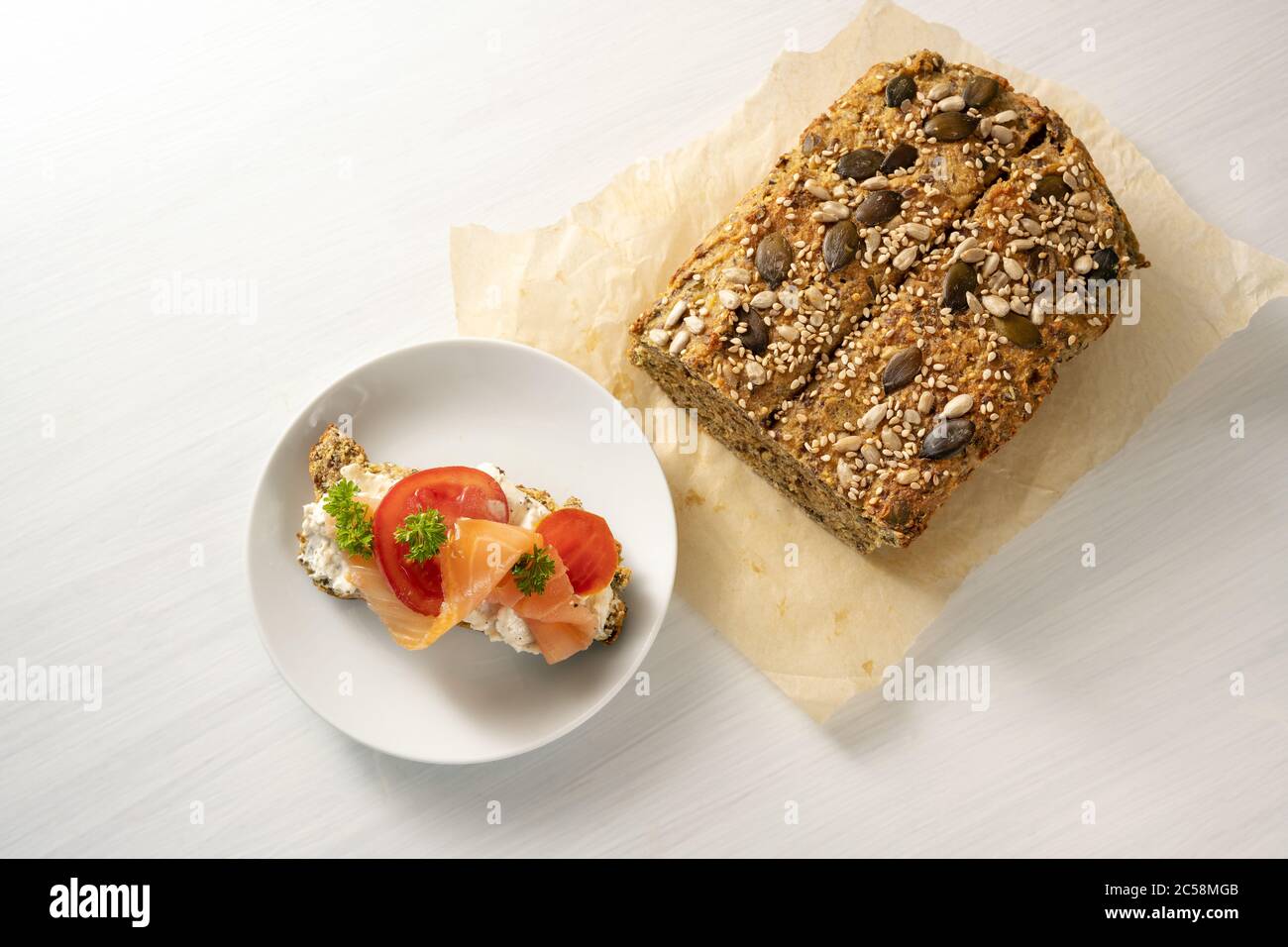 Low carb protein bread with seeds on baking paper and a sandwich with salmon, tomatoes and parsley garnish on a white table, healthy slimming diet, co Stock Photo
