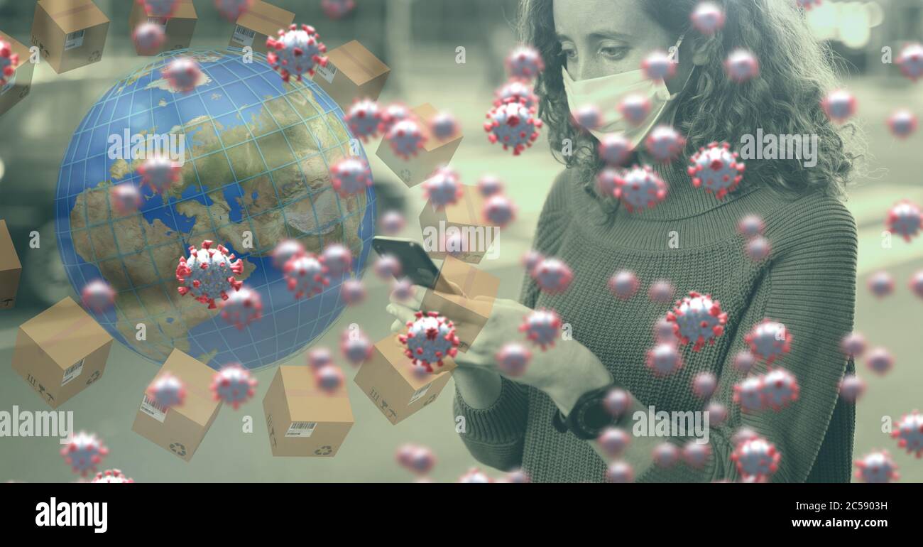 Covid-19 cells and globe surrounded by cardboard boxes against woman wearing face mask using smartph Stock Photo