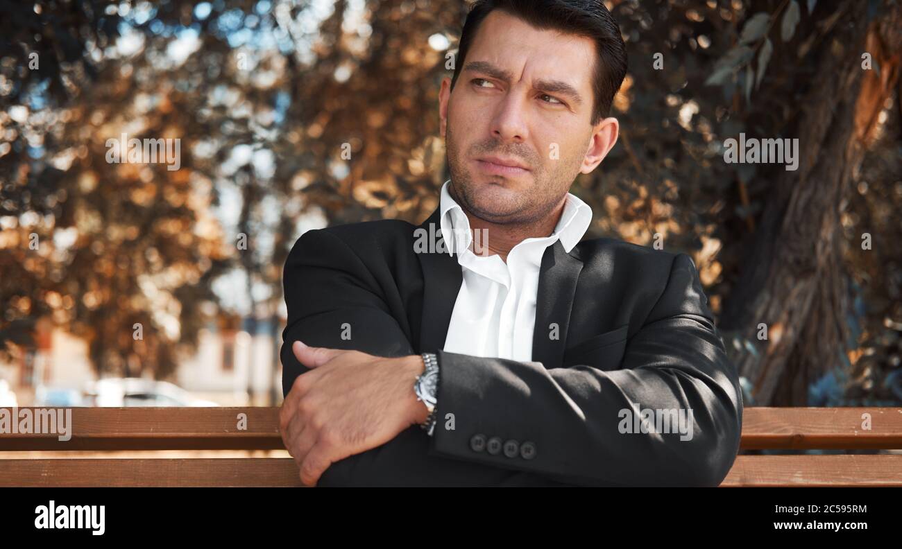 Serious man is looking aside while sitting on a bench in a city park Stock Photo