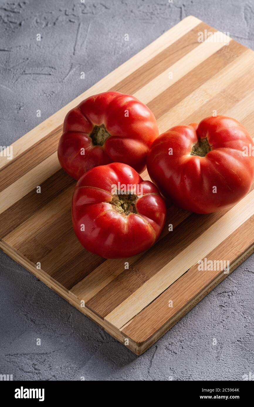 Three pink heirloom tomato vegetables, fresh red ripe tomatoes on wooden cutting board, vegan food, stone concrete background, angle view Stock Photo