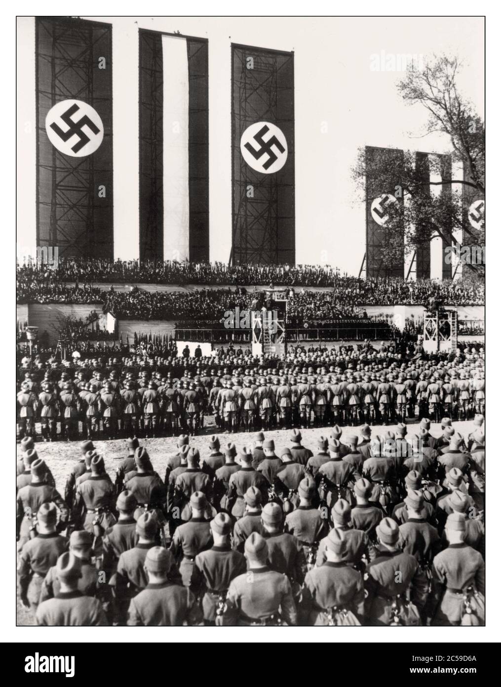 1934 German chancellor Adolf Hitler speaking from an elevated podium, with Nazi Swastika flags hanging as a backdrop behind him, surrounded by German troops at the Tempelhof airport in Berlin, Germany Adolf Hitler delivers his May Day speech in May 1934 Stock Photo