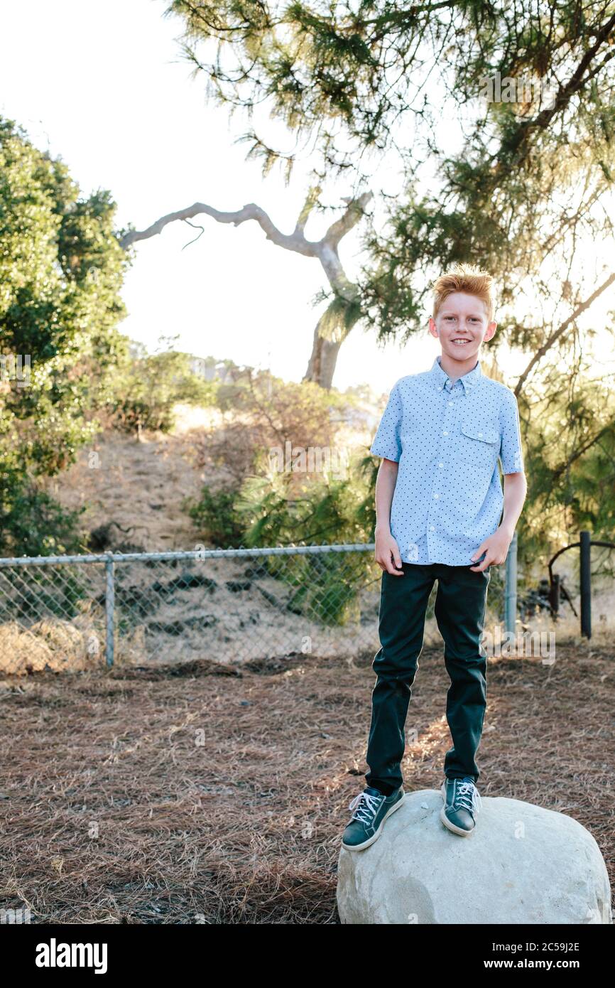 A Red Haired Boy Smiles While Standing On A Large Rock Stock Photo