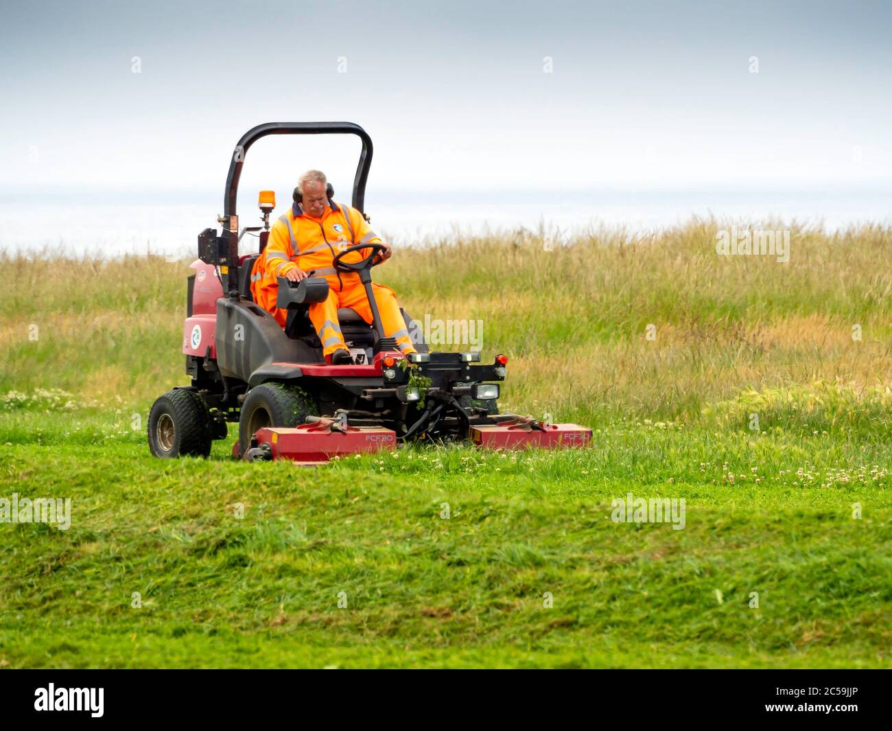 Local Authority Gardener mowing the grass in an open public amenity space on a cliff top in North Yorkshire Stock Photo