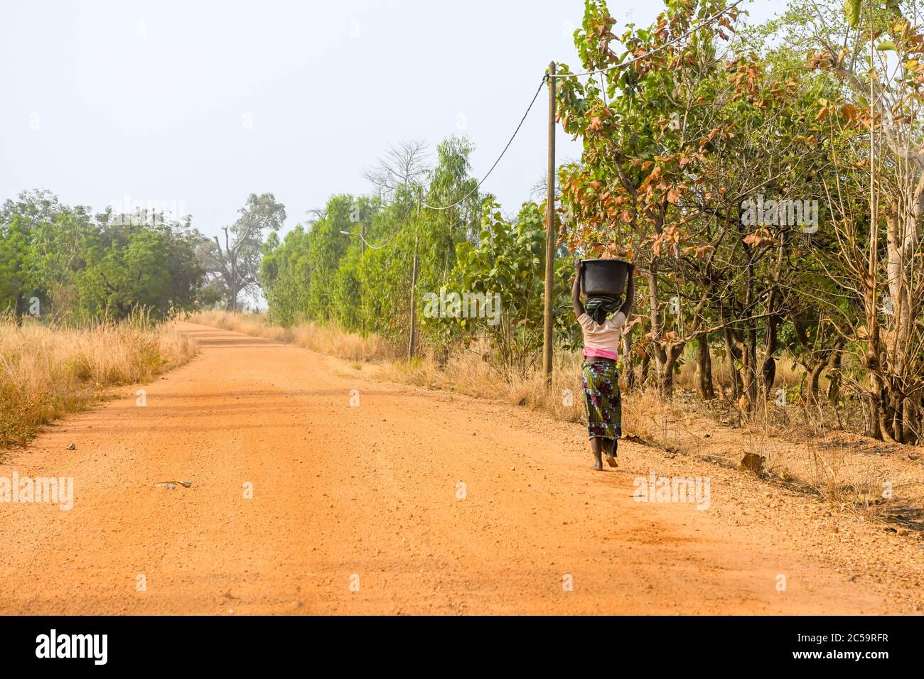 Africa, West Africa, Benin, Natitinqou. A woman walks with a bucket on her head on a dirt road in northern Benin. Stock Photo