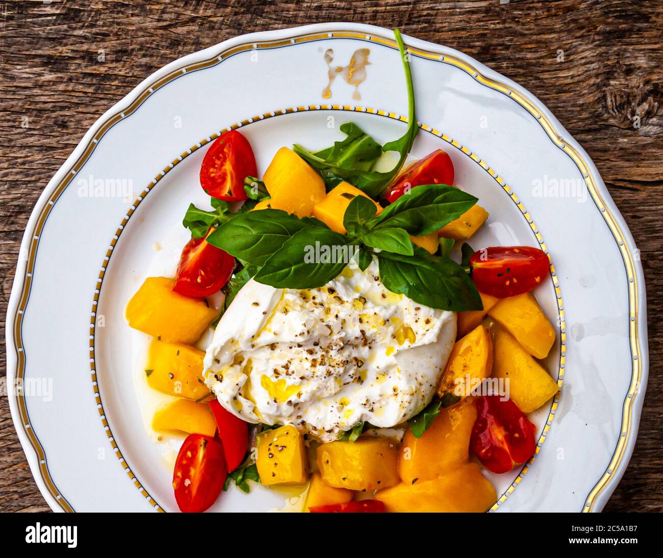 Poodle with a gold rim on the restaurant tableware. Bright yellow in the middle, a wonderful summer dish: burrata with mango and mint salad. Effectively seasoned with olive oil, crunchy sea salt and white pepper Stock Photo