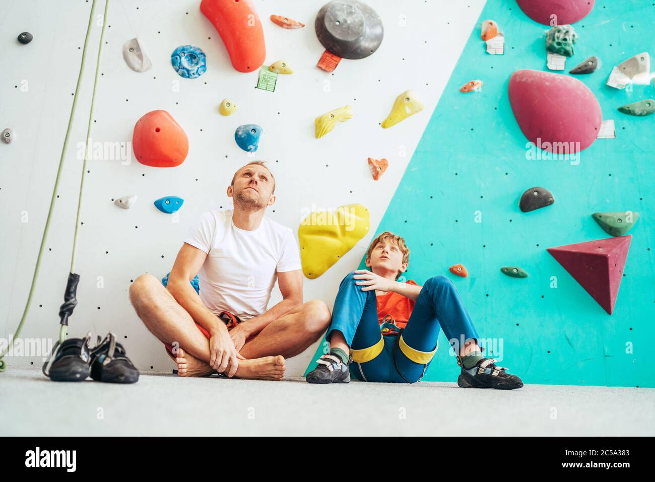 Father and teenage son sitting near the indoor climbing wall. They resting after the active climbing. Happy parenting concept image. Stock Photo