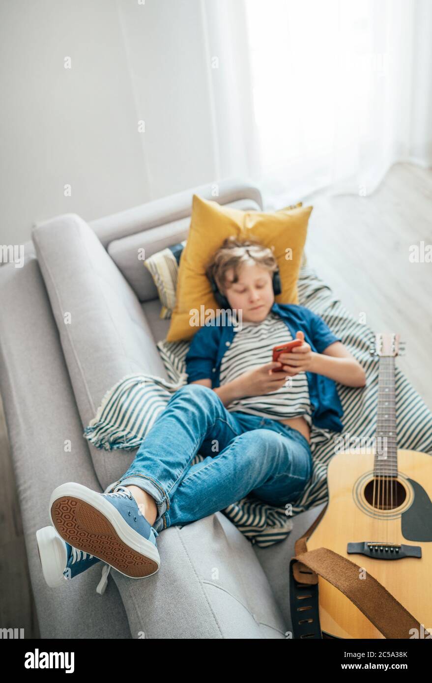 Preteen boy lying with guitar on cozy sofa dressed casual jeans and new sneakers listening to music and chatting using wireless headphones connected w Stock Photo