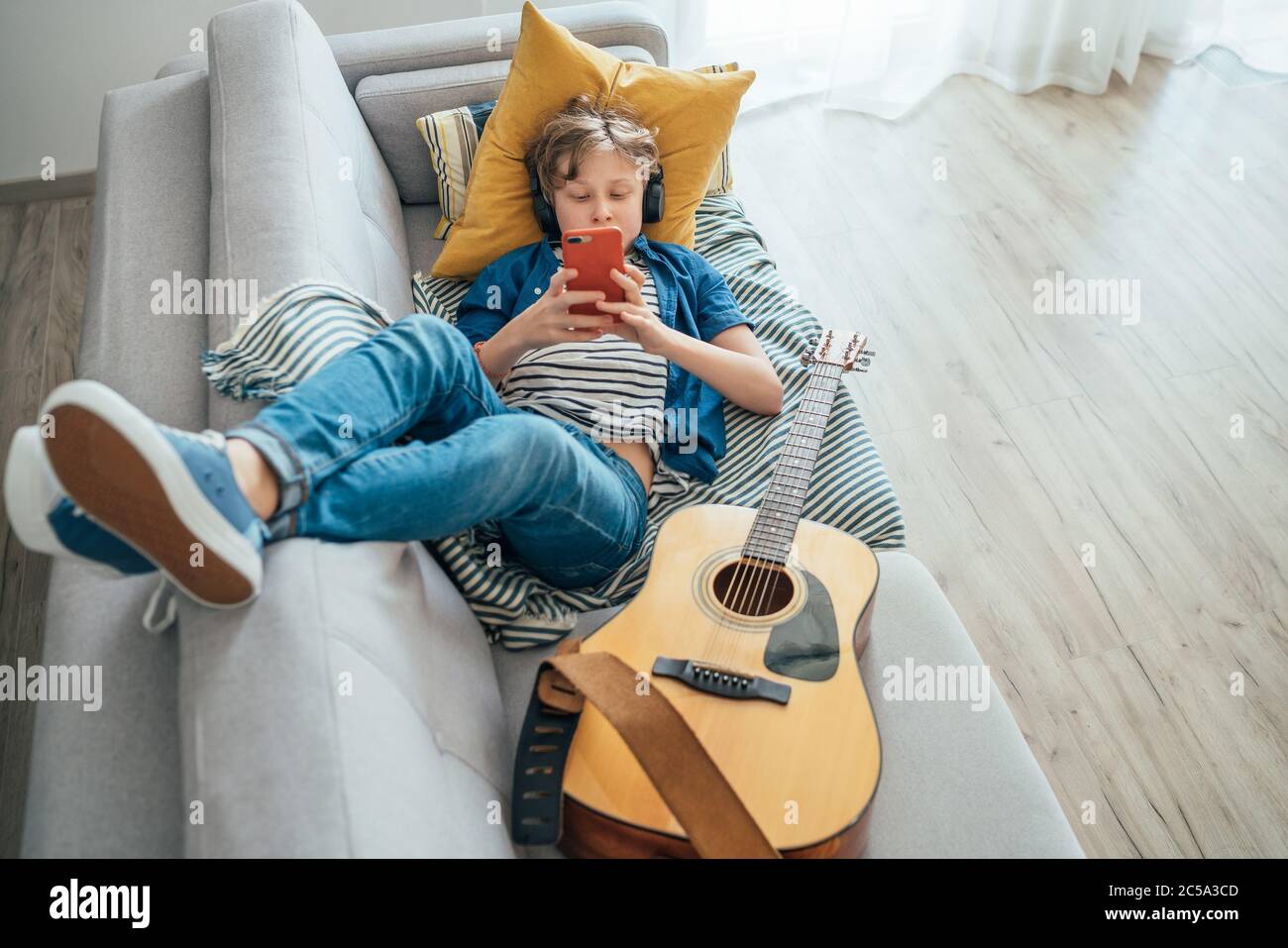 Preteen boy lying with guitar on cozy sofa dressed casual jeans and new sneakers listening to music and chatting using wireless headphones connected w Stock Photo