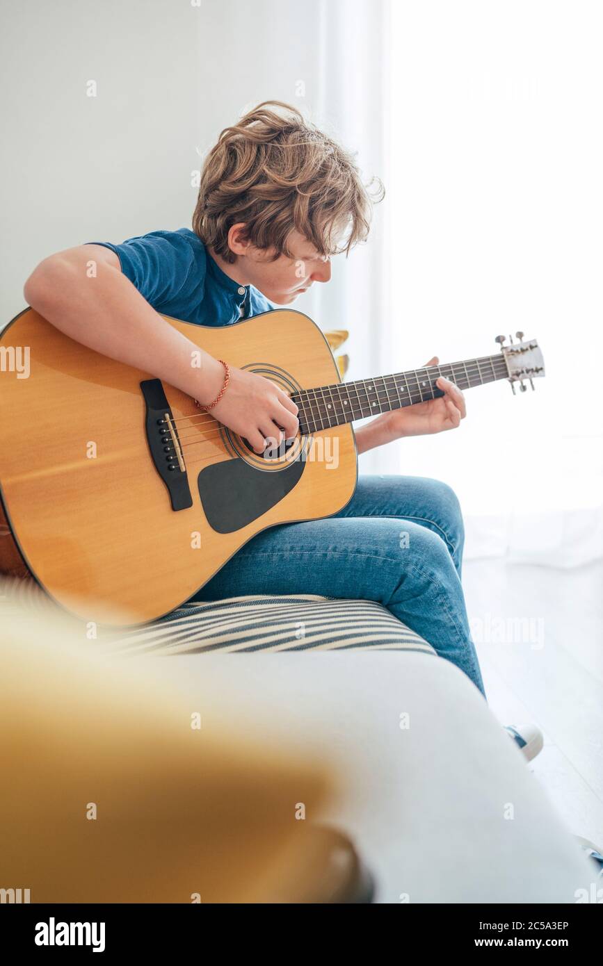 Preteen boy playing acoustic guitar dressed casual jeans and shirt sitting on the cozy sofa at home living room. Music education concept image. Stock Photo