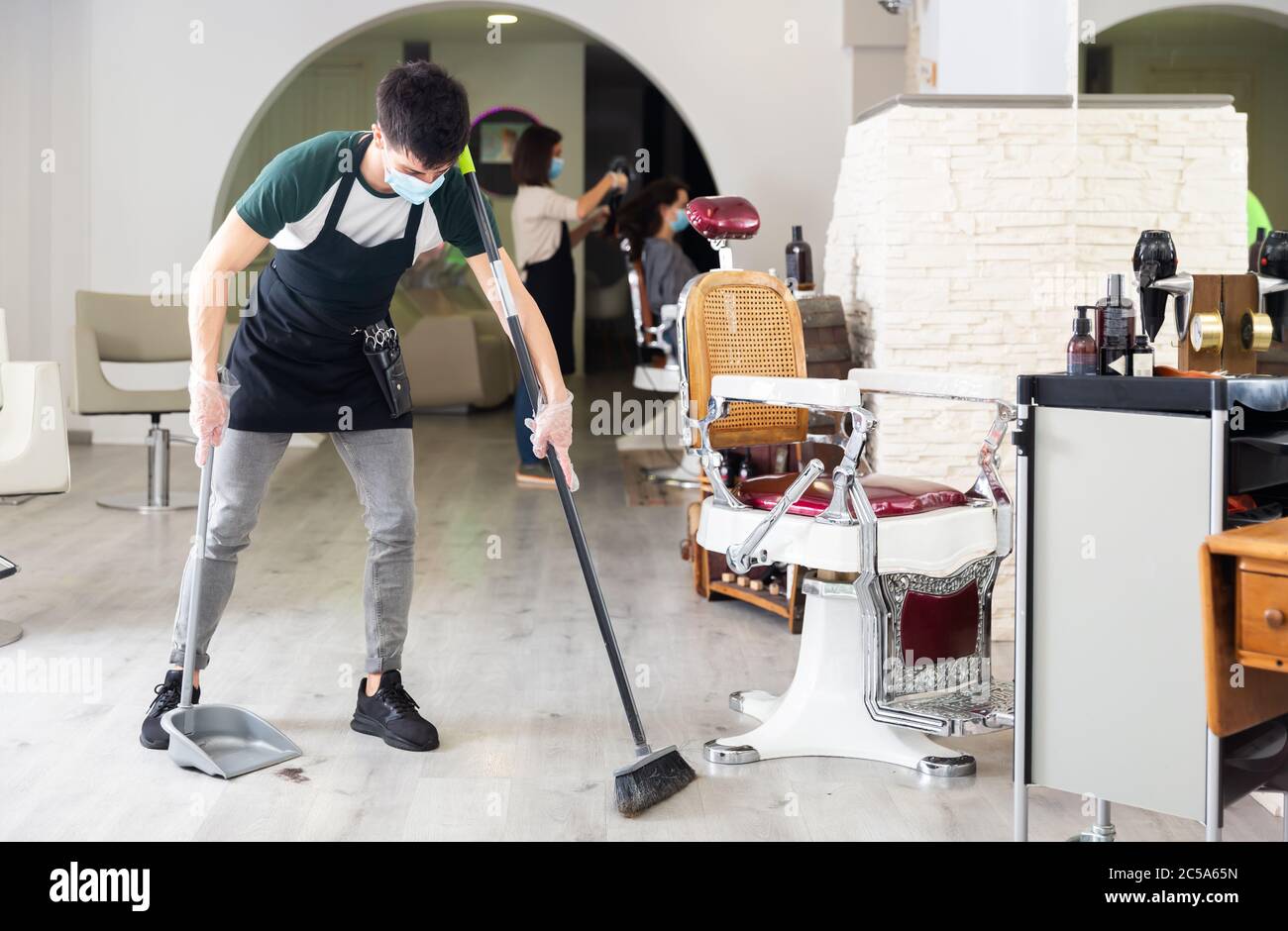 Working process in beauty salon during pandemic situation, man sweeping floor, female hairstyler making hairdo to client on background, all people wea Stock Photo