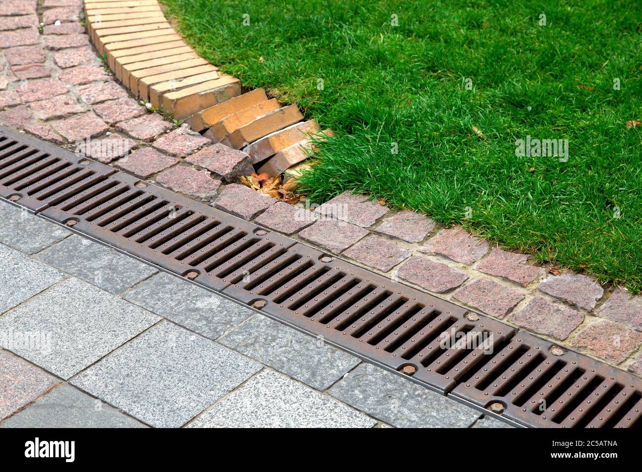 drainage grate of a storm system on a pedestrian sidewalk made of stone tiles in a park near a green lawn. Stock Photo